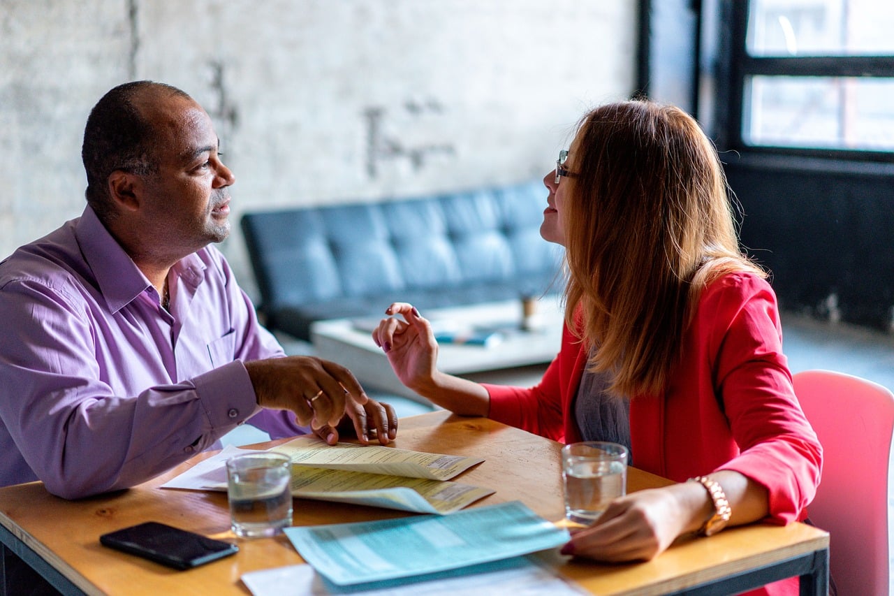 Discussion entre un homme et une femme dans un salon ou un café. Des documents administratifs sont éparpillés sur la table autour de laquelle ils discutent.