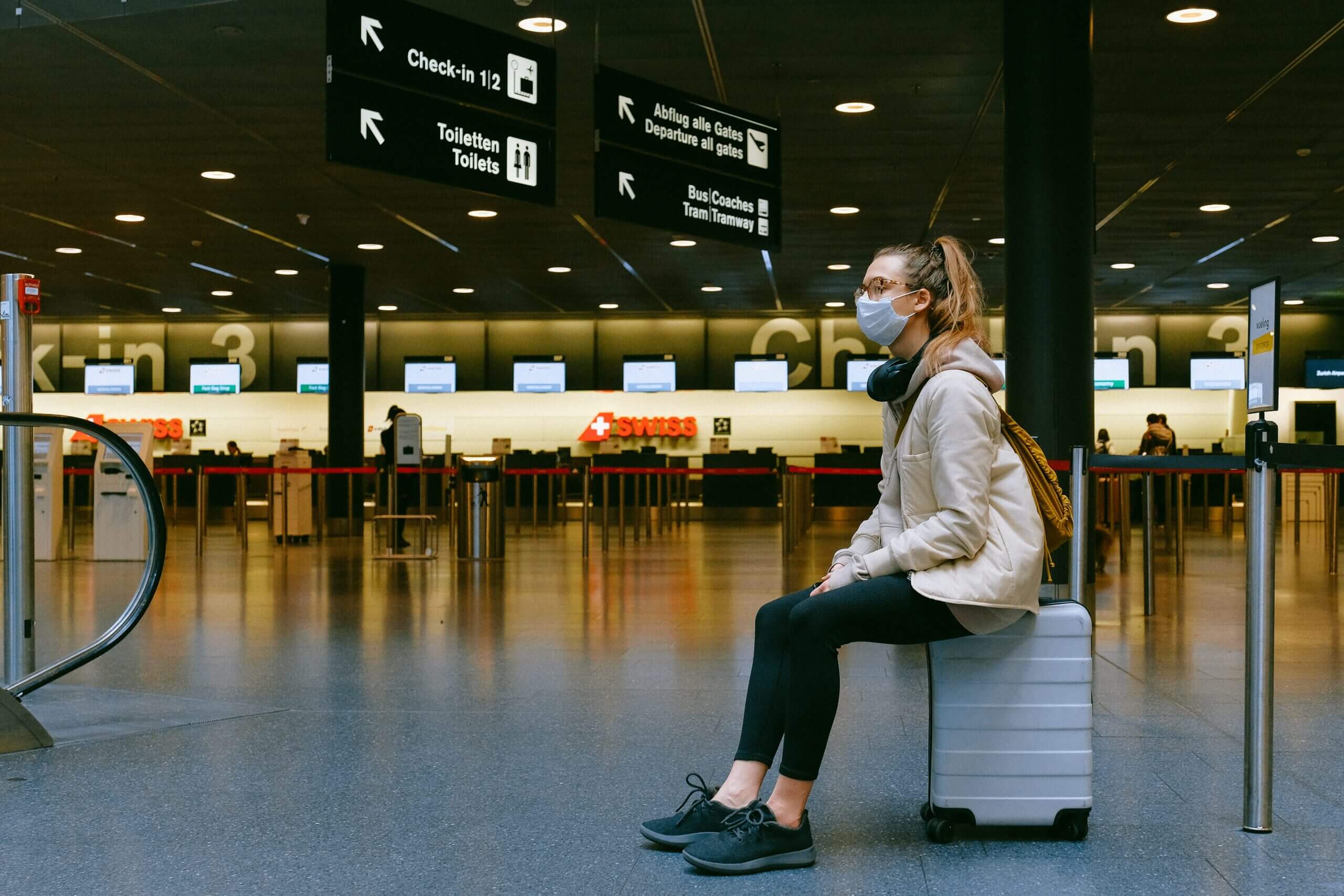 Femme dans un aéroport, assise sur sa valide à roulette, portant un masque chirurgical